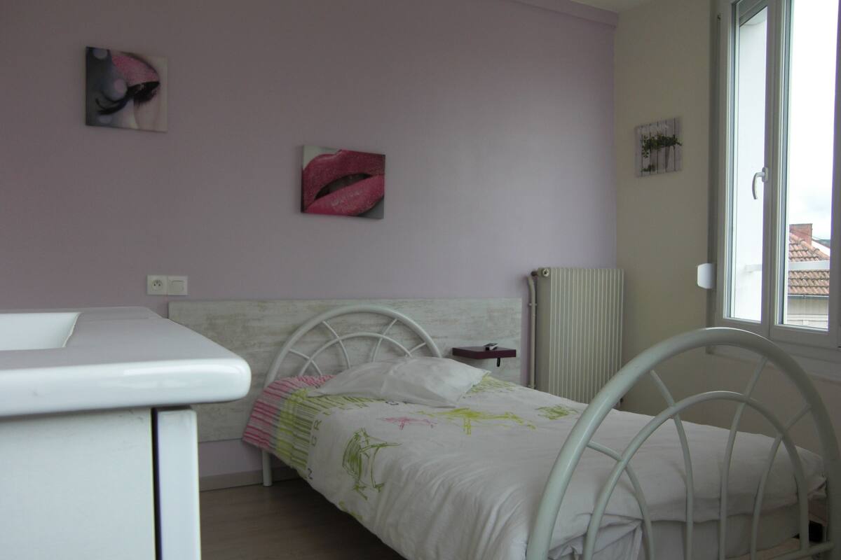A single-bed room with a white metal frame, a bedside table, a radiator, and a window.