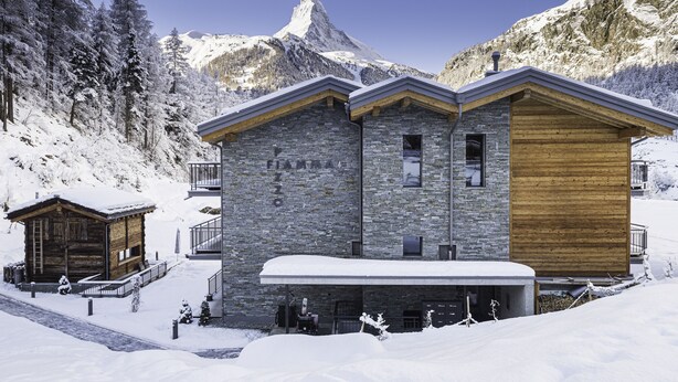 Un chalet à étages, doté d’une façade en pierre et en bois, situé dans une région montagneuse enneigée, avec un sommet imposant en arrière-plan.
