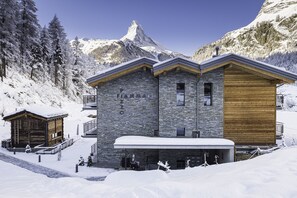 A multi-story lodge with stone and wood exterior, located in a snowy mountainous area with a prominent peak in the background. - Apartment in Chalet Pizzo Fiamma (Zermatt)
