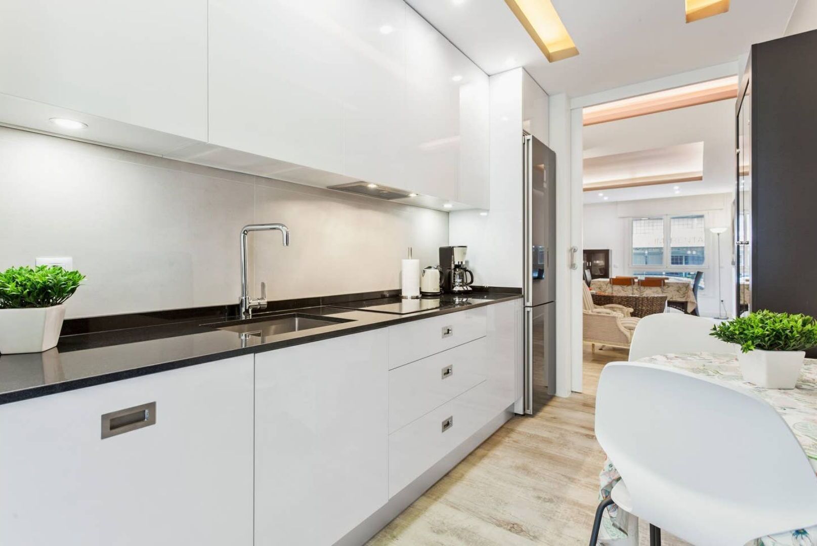 Een moderne keuken met witte kasten, een zwart aanrecht en roestvrijstalen apparatuur. Door een open deur is een eetgedeelte zichtbaar.