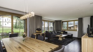 A modern living room with a wooden dining table, dark sofas, and a large window offering a view of a building and greenery. - Chalet Monte Cristo (Zermatt)