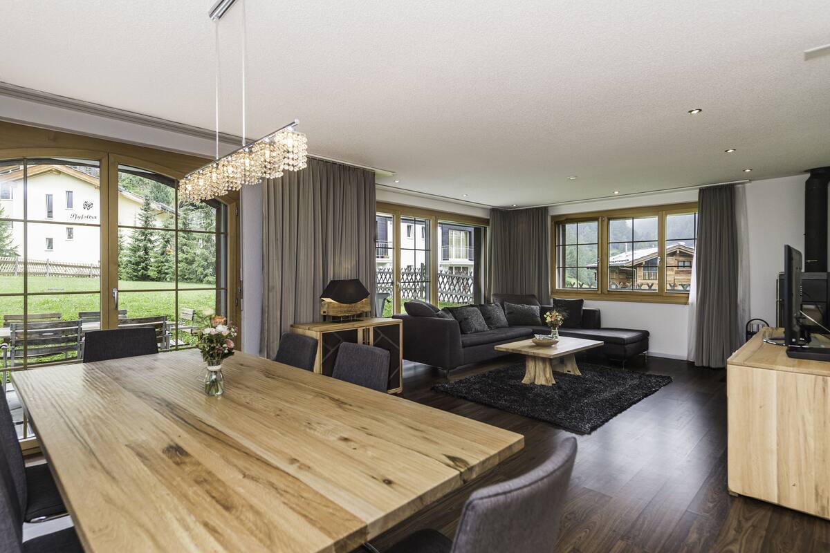 Una sala de estar moderna con una mesa de comedor de madera, sofás oscuros y un amplio ventanal que ofrece vistas a un edificio y zonas verdes.