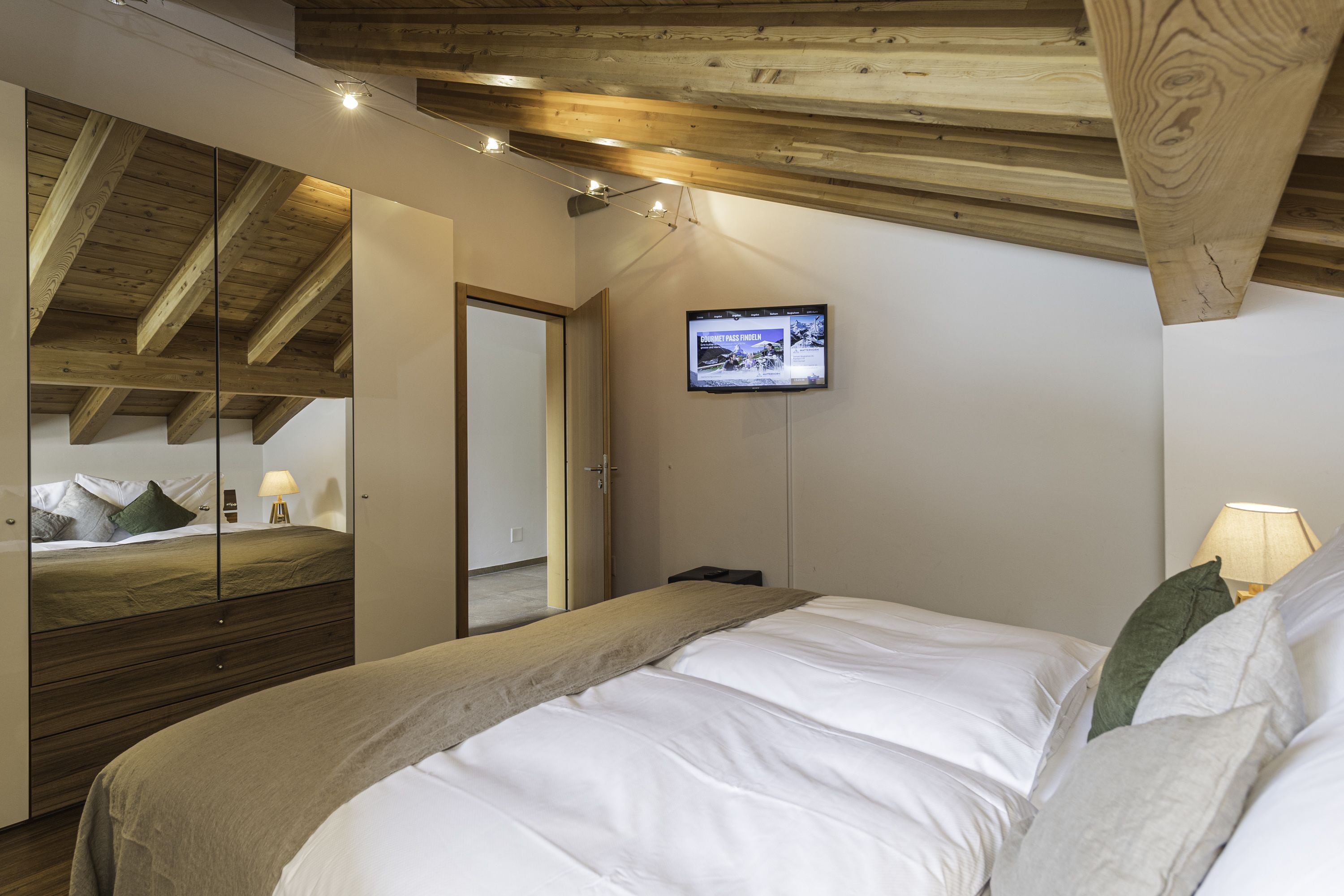 A bedroom with a bed, a television mounted on the wall, and wooden beams on the ceiling.