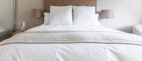 Cama bem arrumada com roupa de cama branca, cabeceira marrom e duas luminárias de cabeceira.