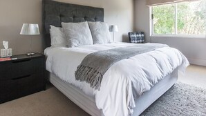 A modern bedroom with a large bed, a dark headboard, and a window with a view of trees. - Houghton Place (Johannesburg)