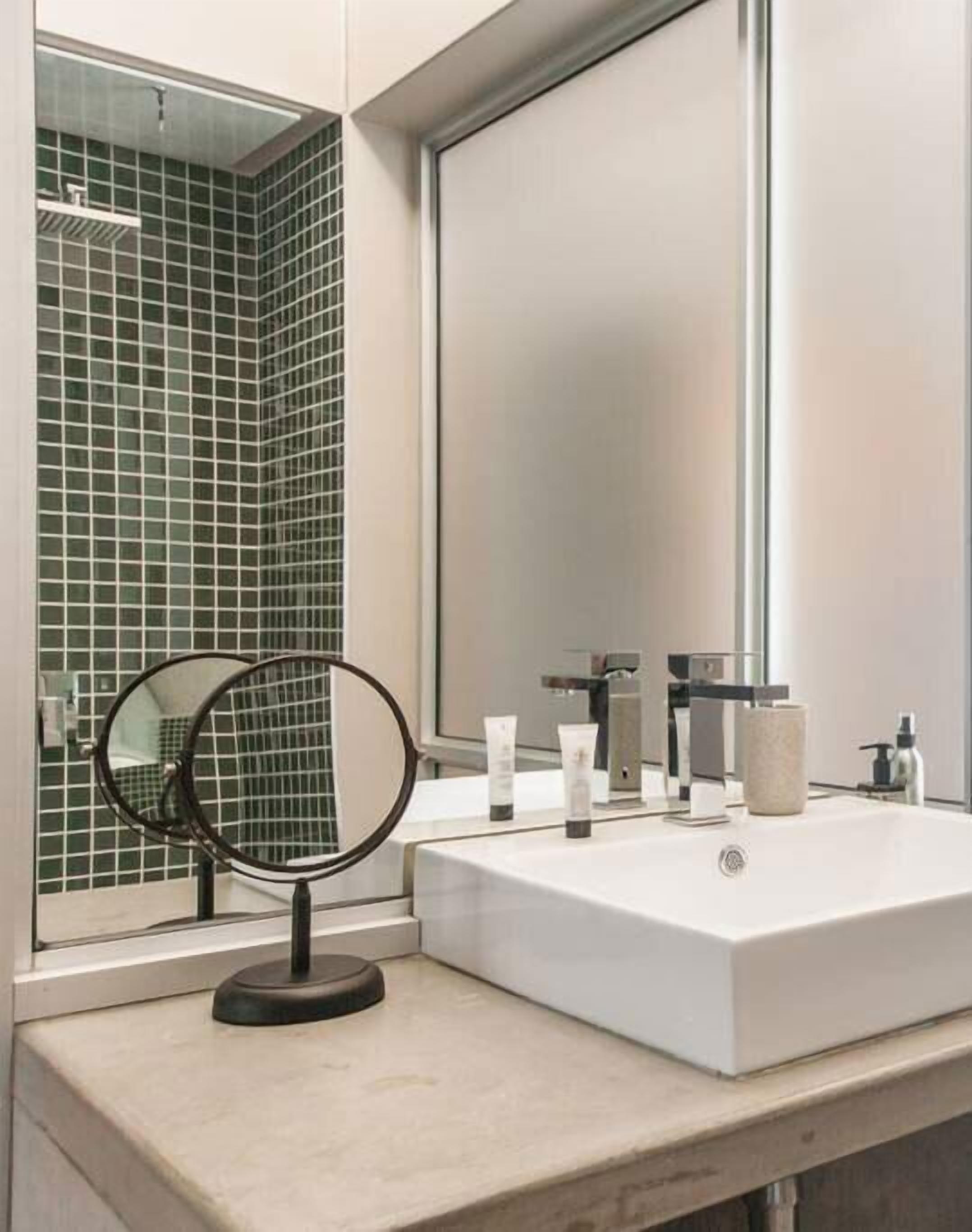Un baño moderno con un espejo grande, un lavamanos blanco y paredes con azulejos verdes.