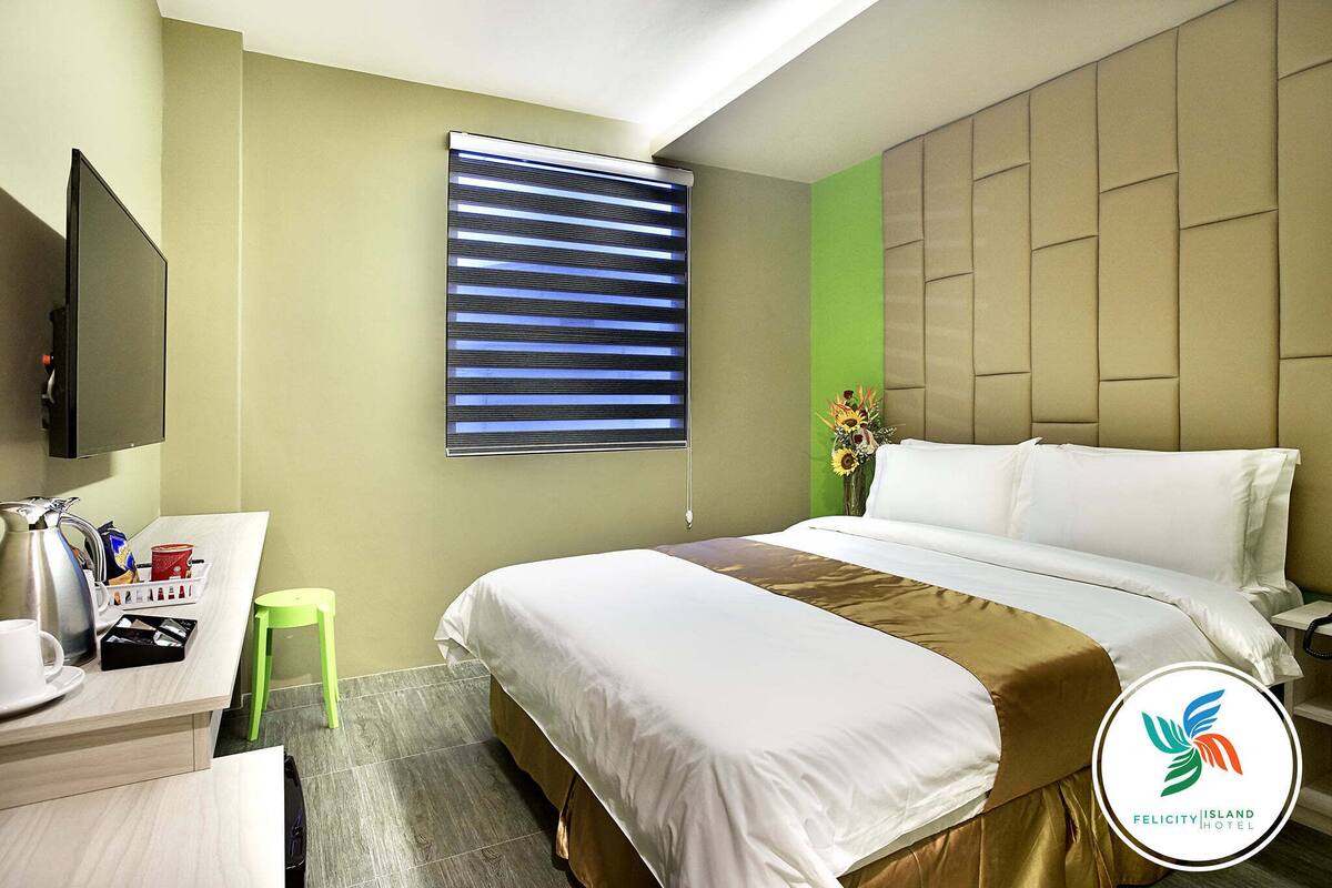 A hotel room with a bed, a TV, a small table with a kettle, a green stool, and a window with blinds.