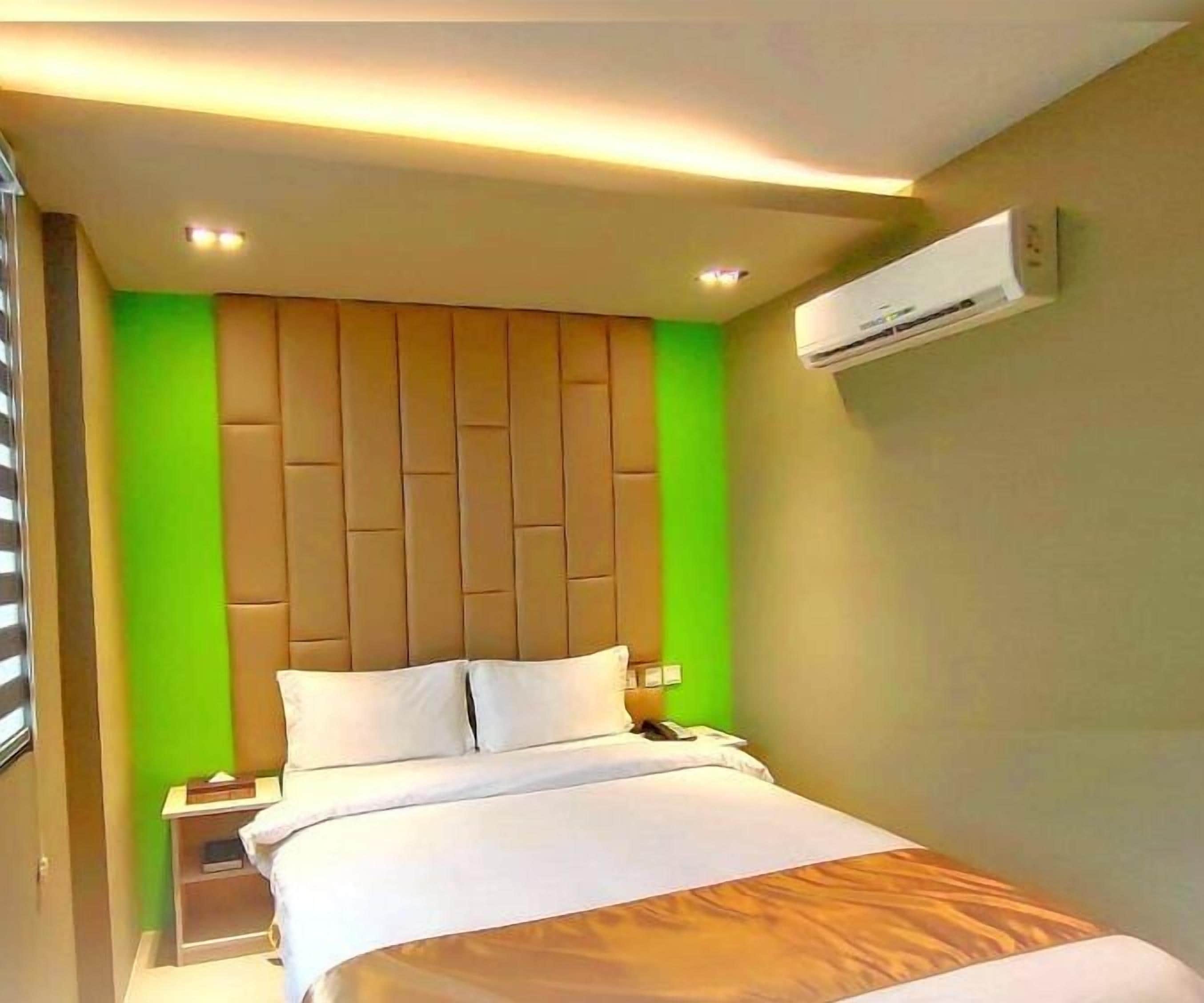 Habitación de hotel con cama, mesita de noche, unidad de aire acondicionado y una pared verde decorativa.