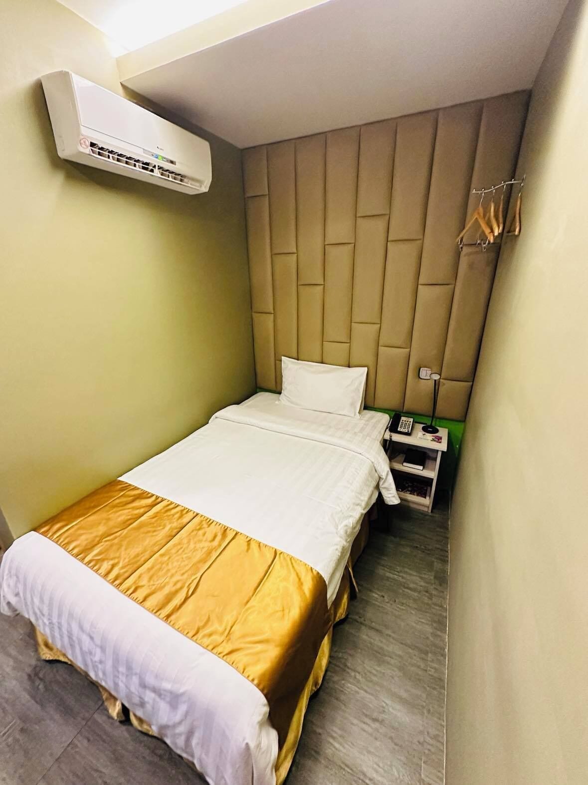 A compact hotel room with a bed, a nightstand, an air conditioning unit, and a small table with a telephone and a book.