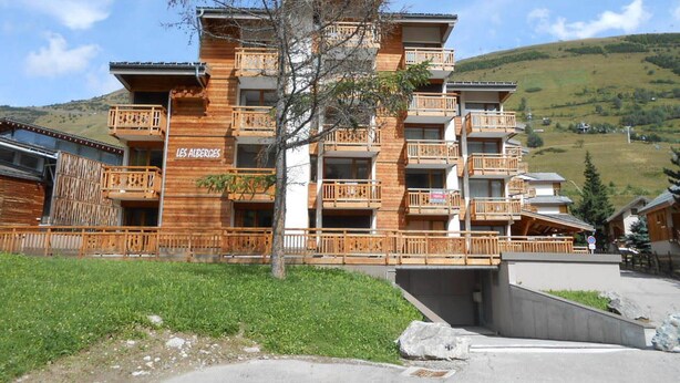 Un edificio de madera de varios pisos con balcones, llamado "LES ALBERGES", ubicado en una zona montañosa con cielo despejado.