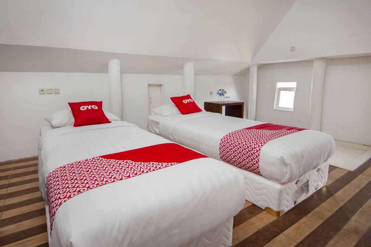 A hotel room with two beds, each with a red and white patterned blanket and a red pillow with "OYO" branding. The room has a wooden floor, white walls, and a small window.