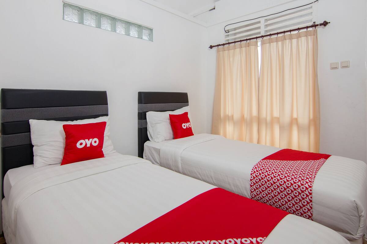 A hotel room with two beds, each with a white bedspread and red pillows, and a beige curtain.