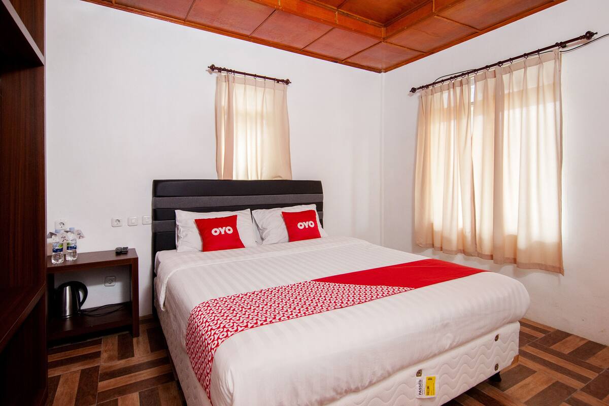 A hotel room with a bed, a nightstand, a red and white patterned bedspread, and two windows with sheer curtains.