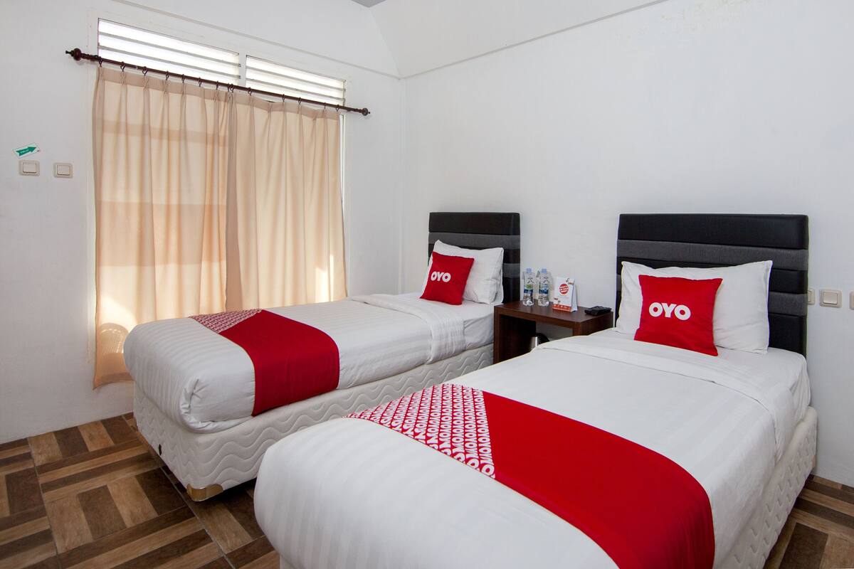 A hotel room with two beds, each with a red and white bedspread, a nightstand with a bottle of water, and a window with beige curtains.