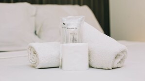 A neatly made bed with white linens, a rolled towel, a cotton swab, and a small bottle labeled "Essential Kit."
