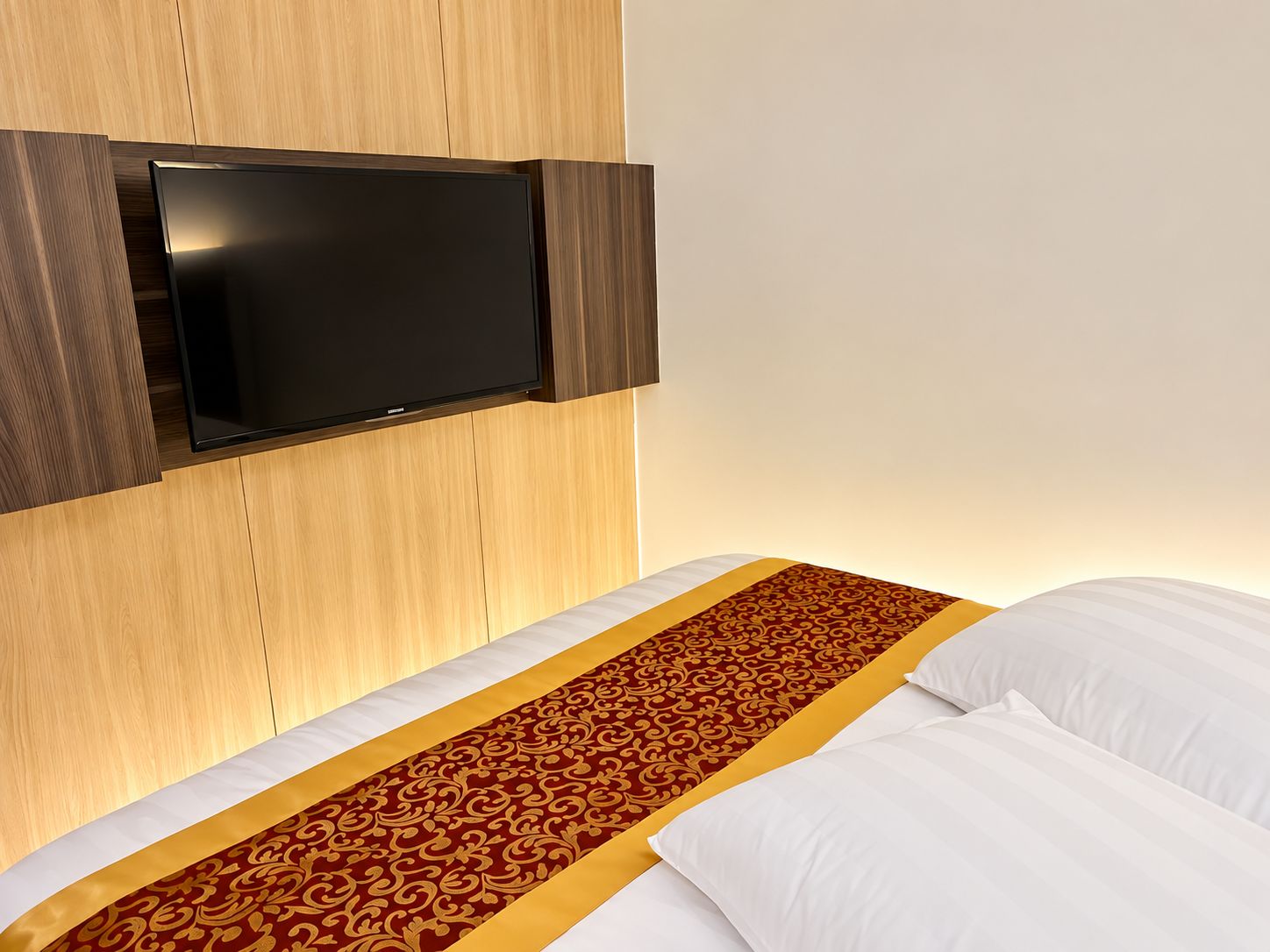 A hotel room with a bed, a flat-screen TV mounted on the wall, and a patterned bed runner.
