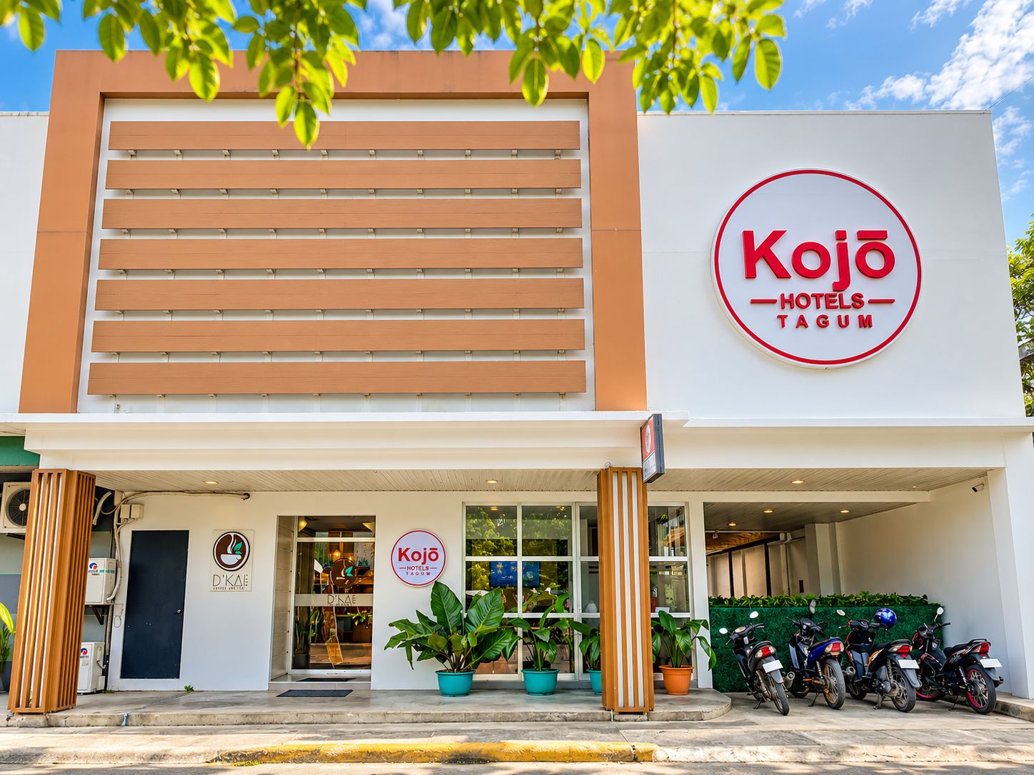A hotel named Kojo Hotels- Tagum with a modern facade, a visible entrance, and a parking area with motorcycles.