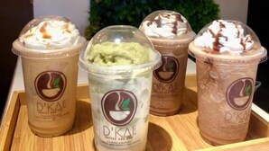 A tray with four D'KAE iced drinks, one with a green matcha flavor, and a green wall with plants in the background.