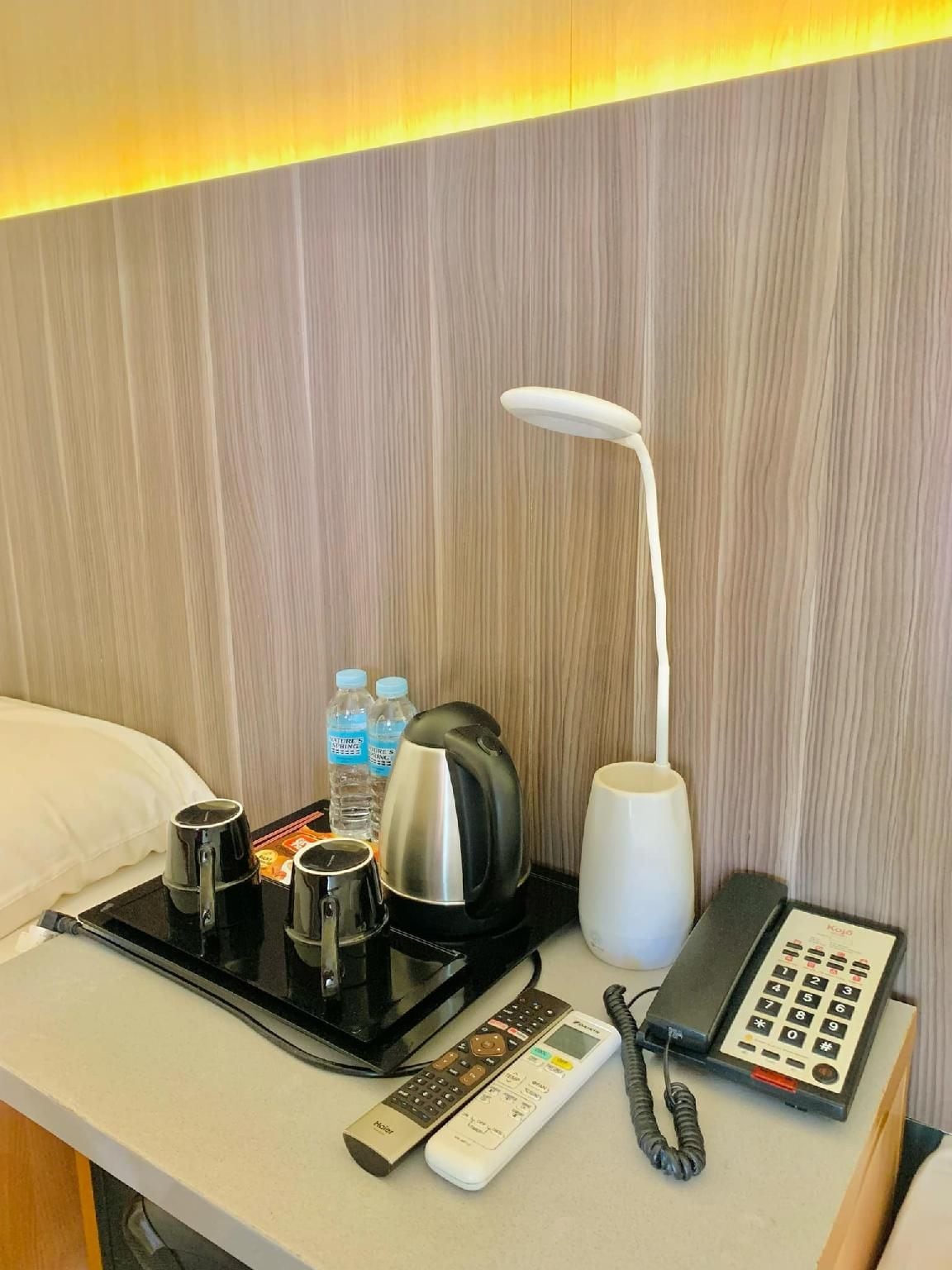 A hotel room with a bed, a bedside table with a kettle, a coffee maker, a water bottle, a remote control, and a telephone.