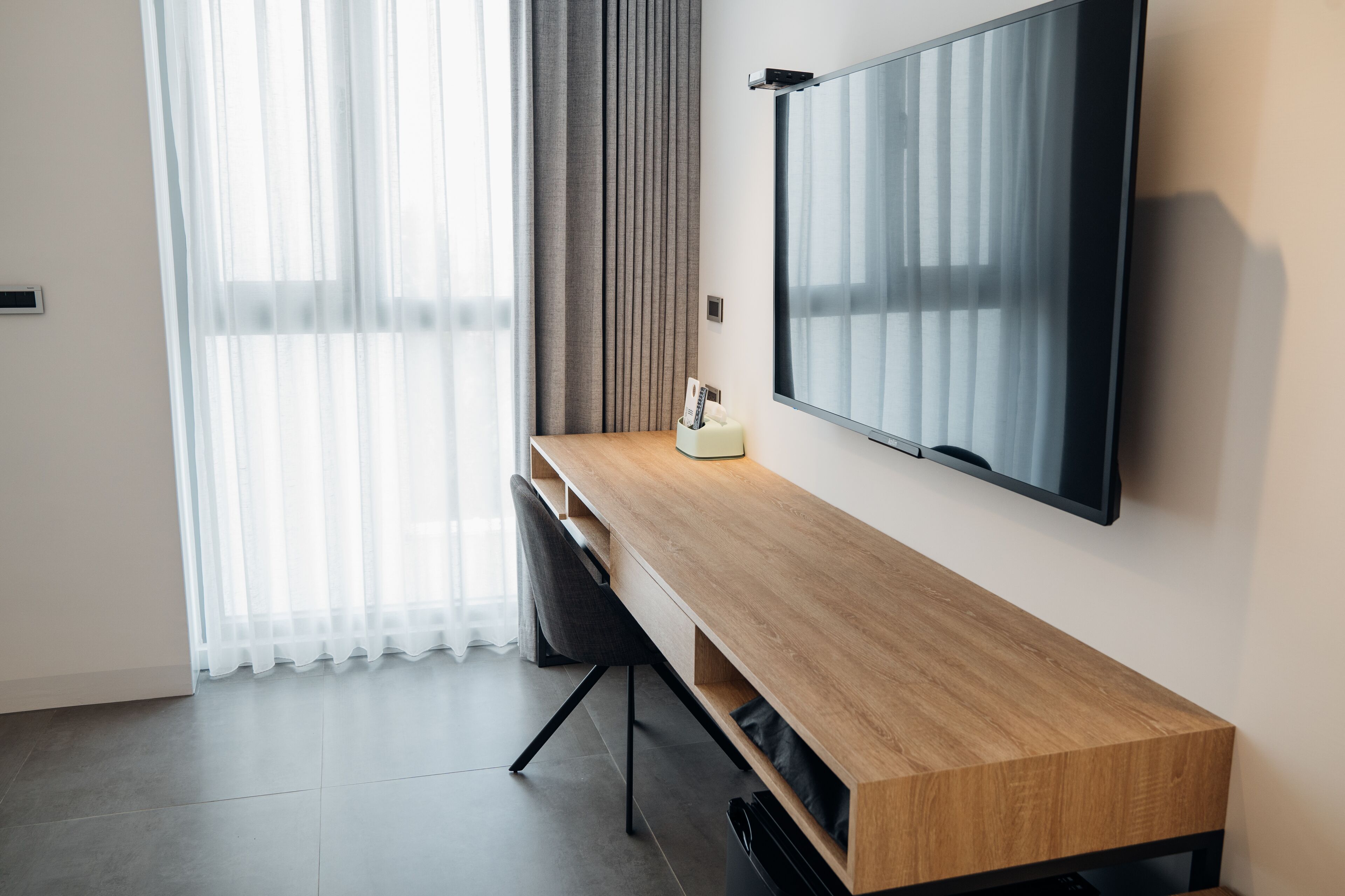 A modern hotel room with a wooden desk, a flat-screen TV, a chair, and a window with sheer curtains.