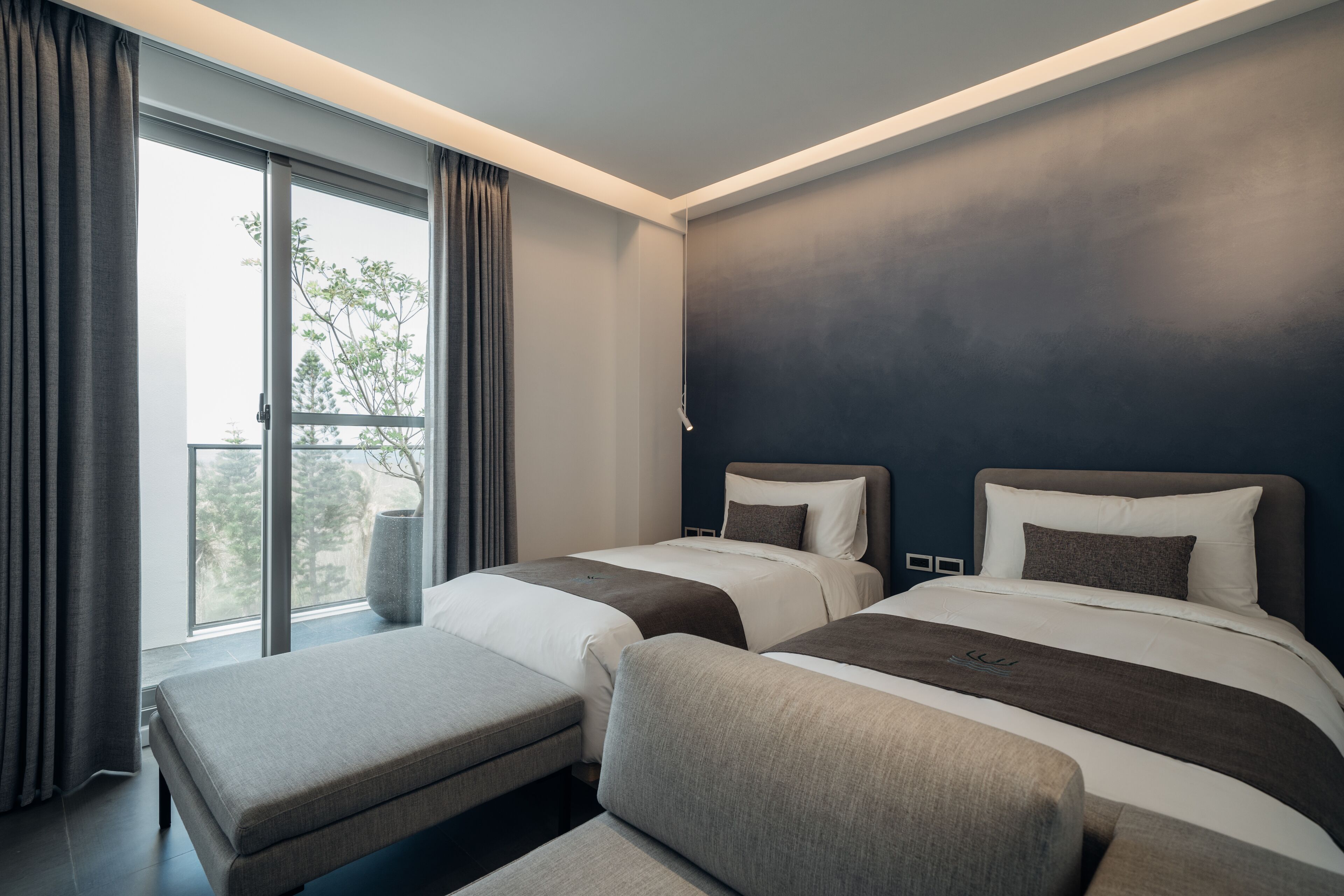 Ein modernes Hotelzimmer mit zwei Betten, einem großen Fenster inklusive Vorhängen, einer Bank und Blick auf Bäume.