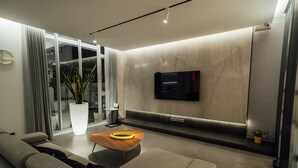 Una sala de estar moderna con un sofá, una mesa de centro, un televisor montado en la pared y un amplio ventanal con cortinas.