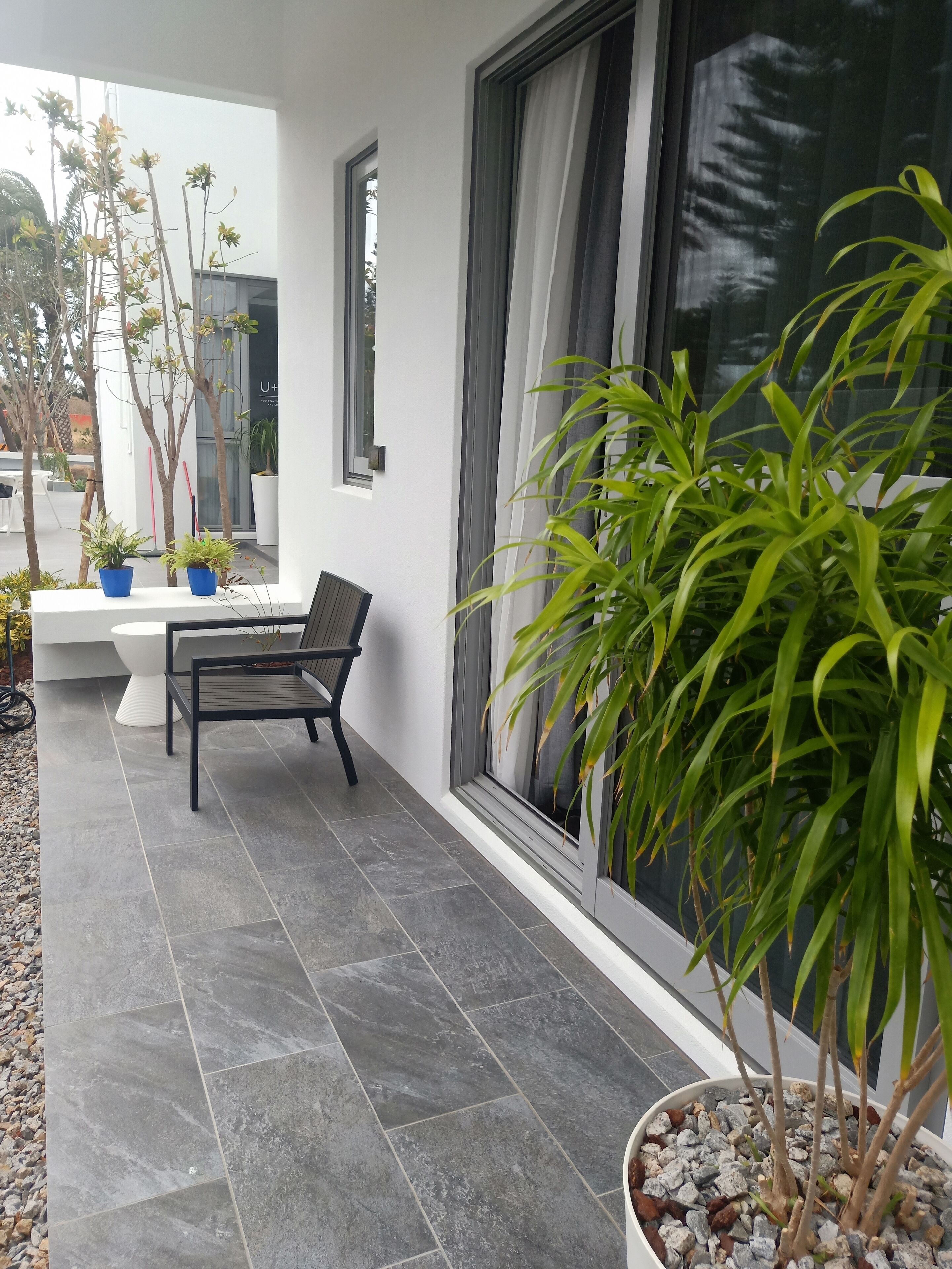 Uma área externa moderna com área de estar, um banco e plantas em vasos, um terraço com ladrilhos e uma janela grande com cortinas.