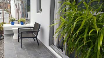 Uma área externa moderna com área de estar, um banco e plantas em vasos, um terraço com ladrilhos e uma janela grande com cortinas.