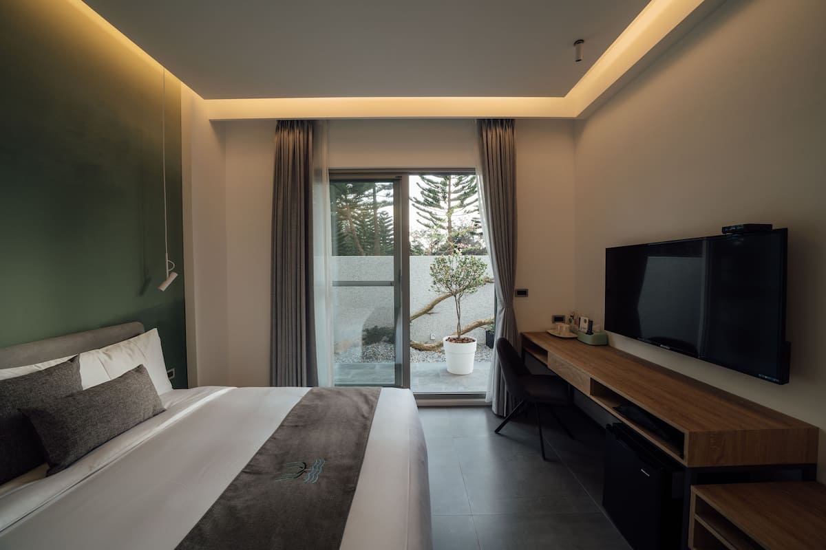 A modern hotel room with a large bed, a TV, a desk, and a view of a tree through the glass door.