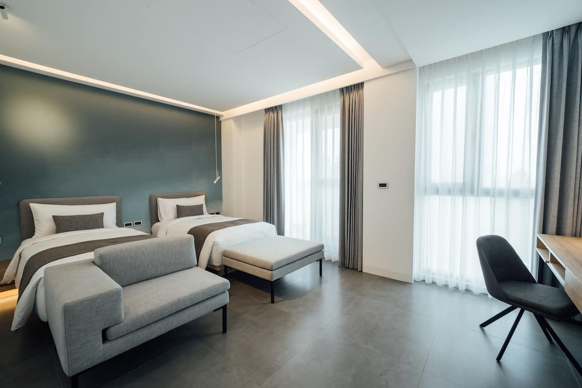 A modern hotel room with two beds, a sofa, a desk, and large windows with curtains.