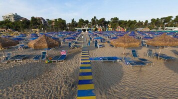 Una playa con filas de tumbonas y sombrillas de paja, un camino central y edificios al fondo.