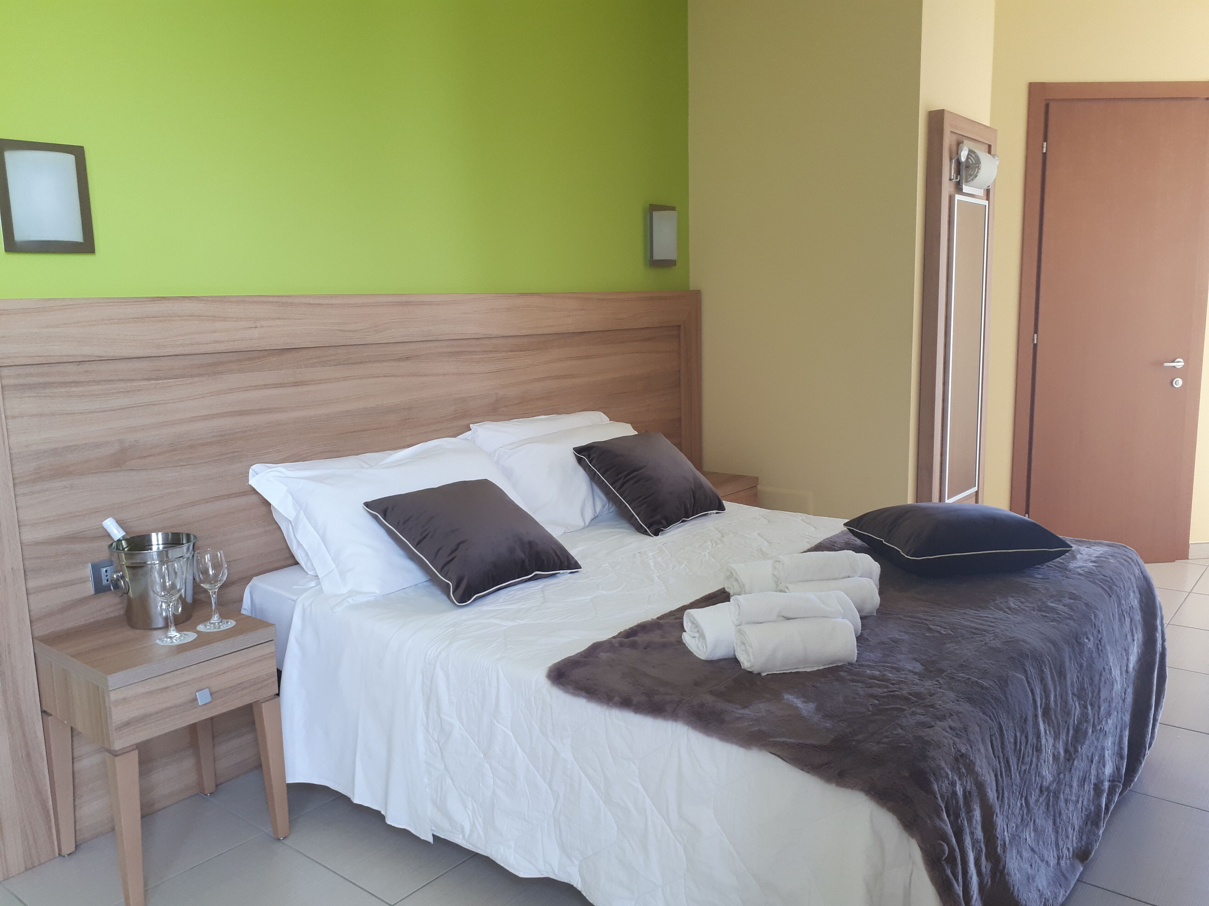 A neatly made bed with white linens and two dark pillows, a wooden nightstand with a glass of water and a wine glass, and a brown furry throw blanket.