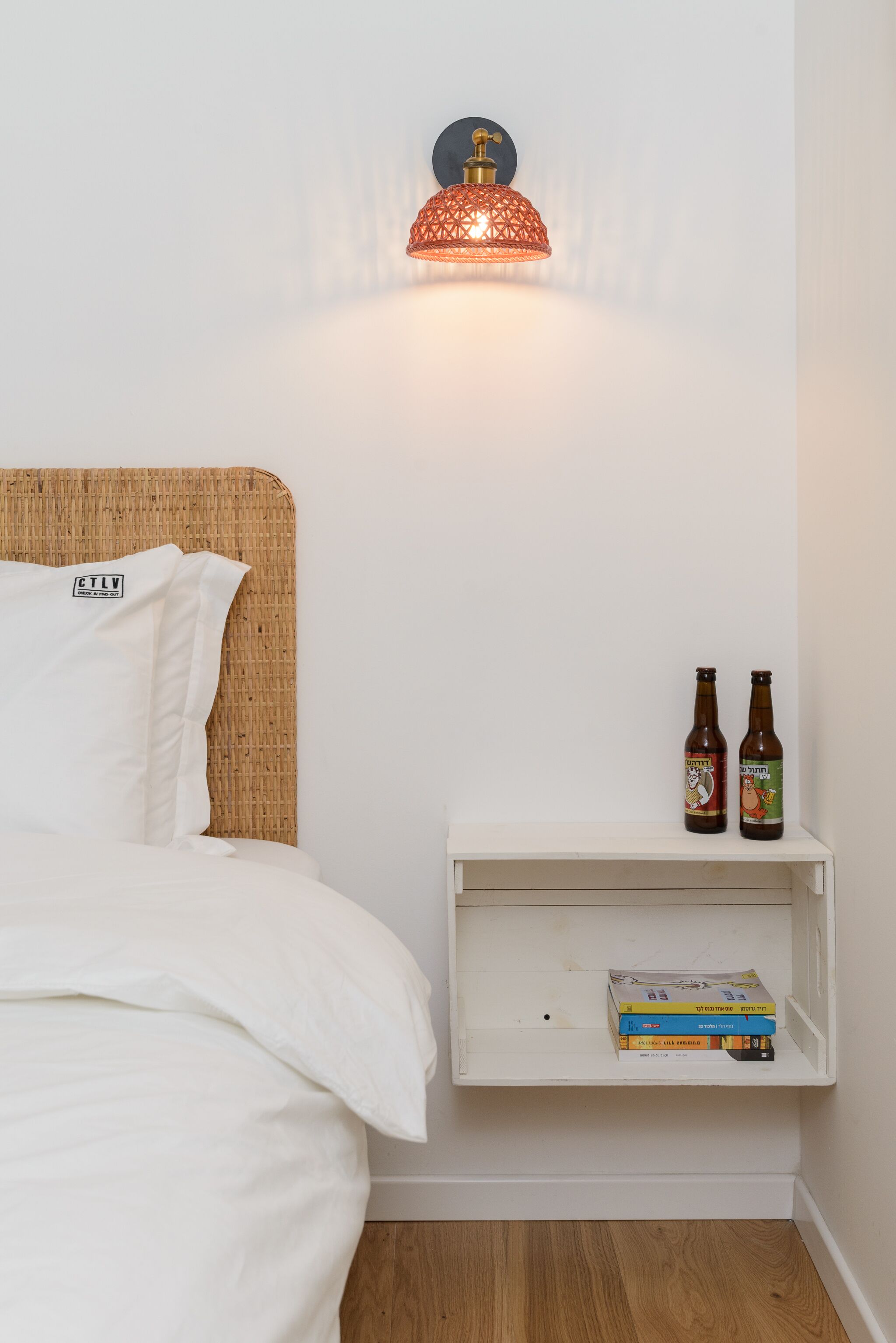 A neatly made bed with white bedding, a wicker headboard, and two bottles of beer on a white nightstand.