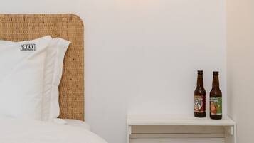 A neatly made bed with white bedding, a wicker headboard, and two bottles of beer on a white nightstand.