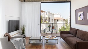 Un salon moderne avec un canapé, une table basse et un téléviseur. On aperçoit un balcon aménagé avec du mobilier d’extérieur.