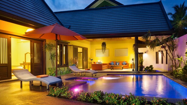 Zona de piscina con sillones de descanso, una sombrilla roja e interiores bien iluminados.