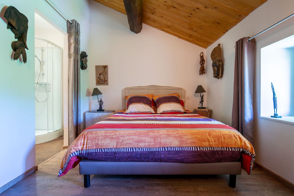 Une chambre avec un grand lit, des poutres apparentes en bois au plafond, un espace douche et des œuvres d’art murales.