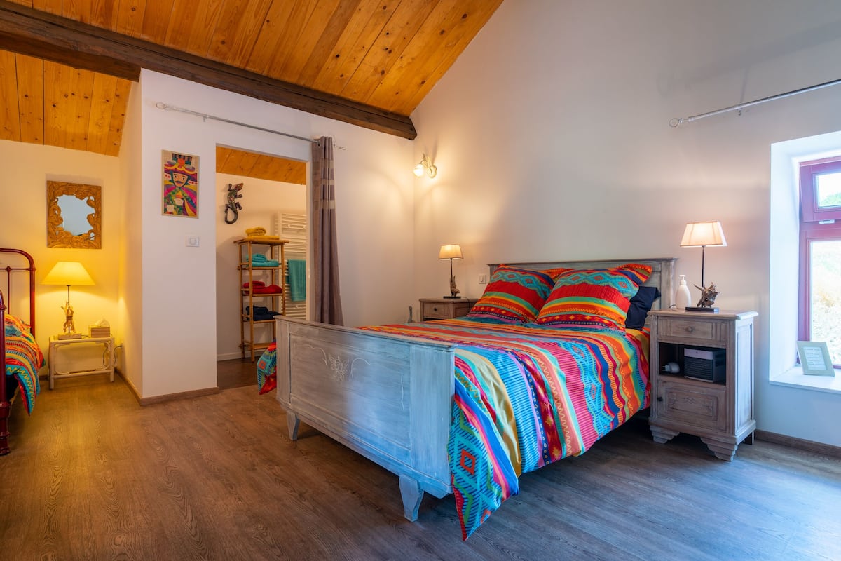 Une chambre avec un plafond en bois, un lit avec une literie aux couleurs vives, une table de chevet avec une lampe et une fenêtre donnant sur un beau paysage.