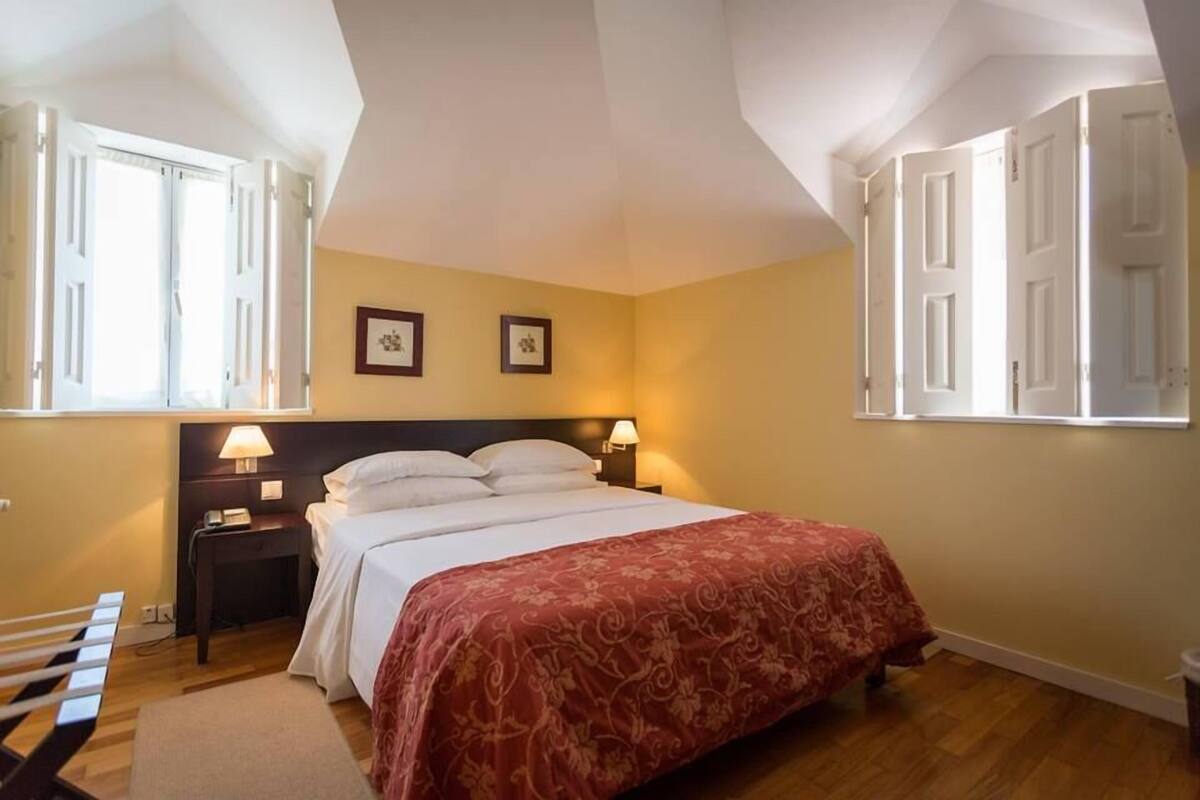 Une chambre à coucher avec un lit, deux lampes de chevet, une table de chevet, une fenêtre avec des volets et un cadre photo accroché au mur.
