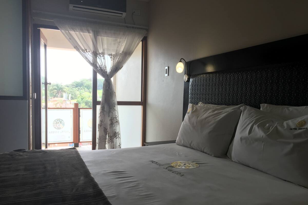 A hotel room with a bed, a headboard, a window with a view of greenery, and a curtain.