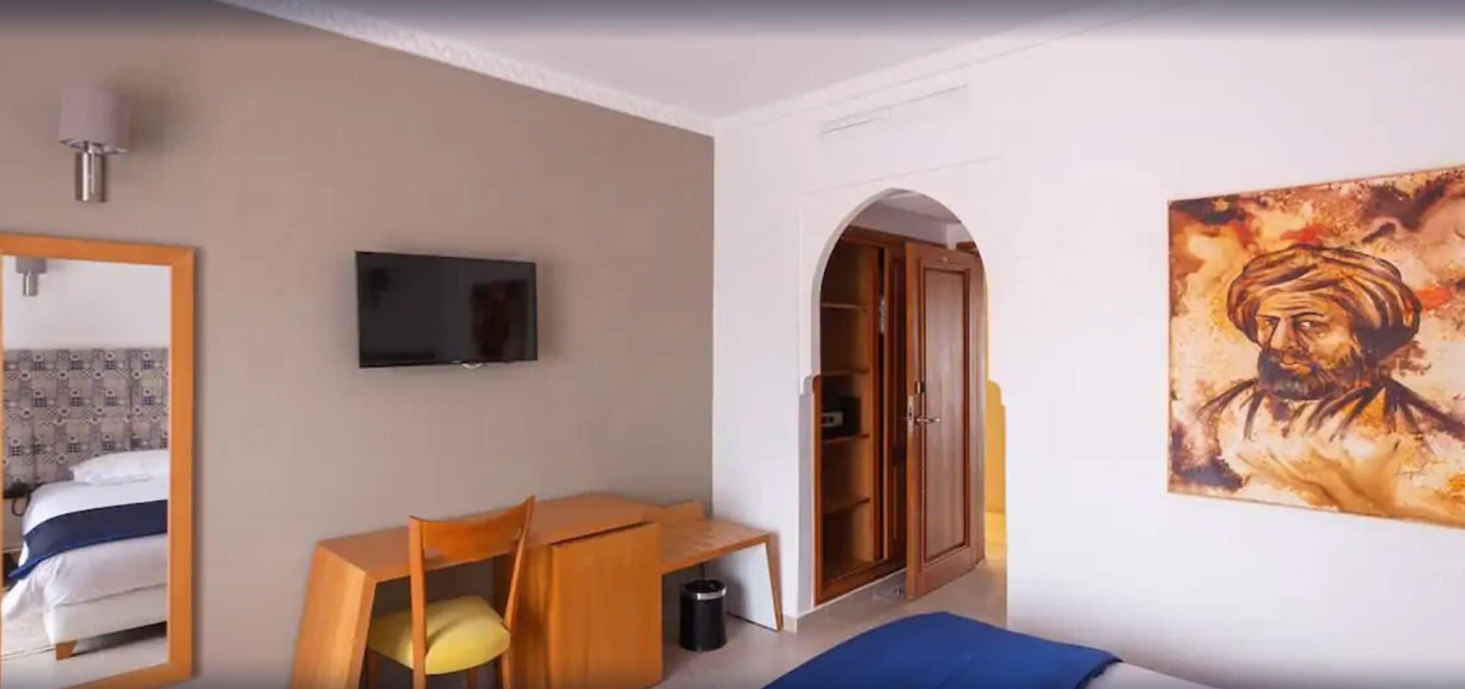Quarto de hotel com cama, escrivaninha, cadeira, televisão, guarda-roupa e um quadro na parede.