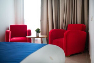 Hotellihuone, jossa on sänky, kaksi punaista nojatuolia, pieni pyöreä pöytä, jossa on kasvi, ja näkymä ulos ikkunasta verhojen läpi.