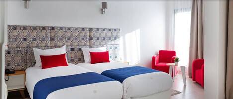 A hotel room with two beds, a red chair, a small table with a plant, and a patterned headboard.