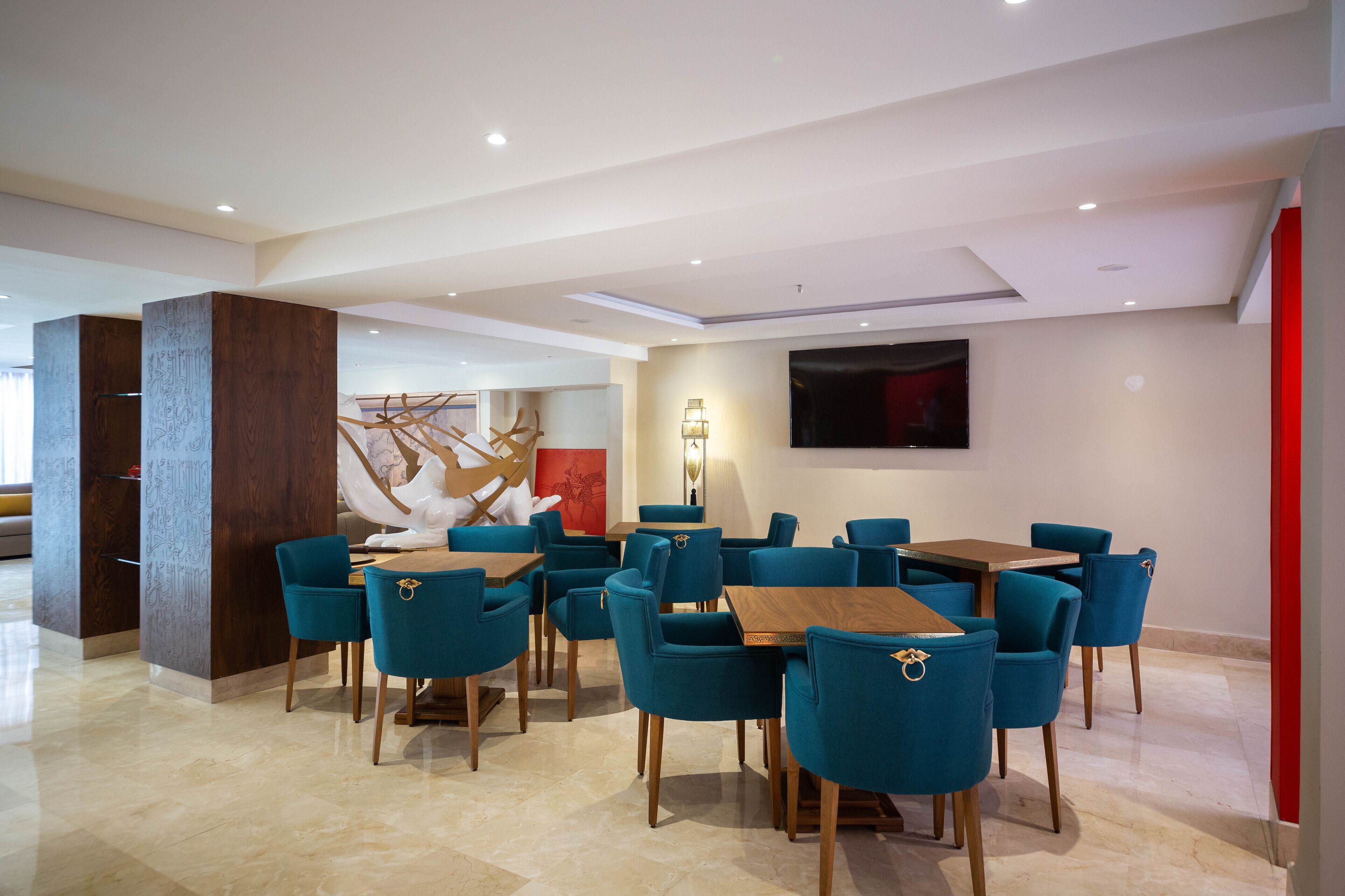 A modern dining area with teal chairs and wooden tables, a large abstract art piece, and a flat-screen TV on the wall.