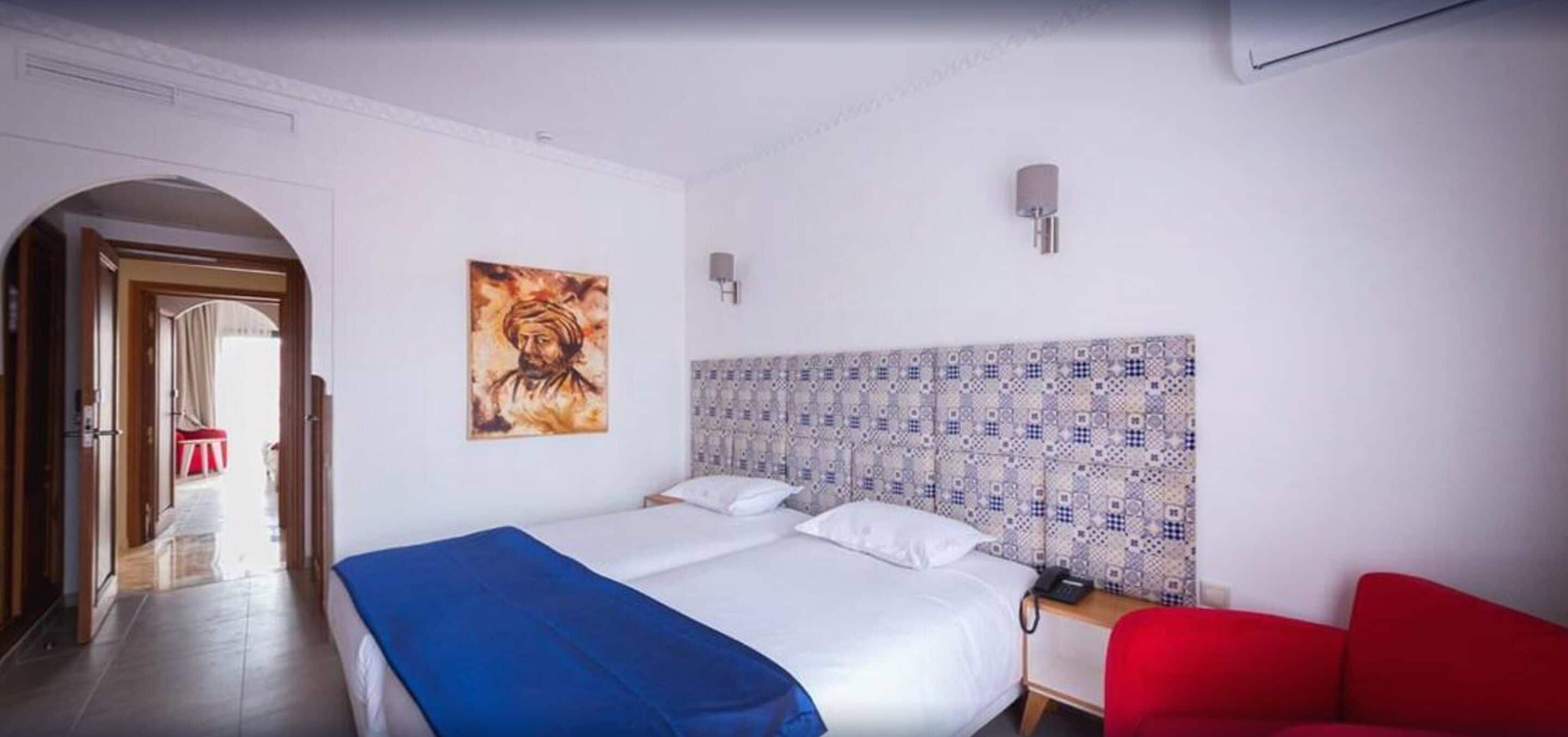 Quarto de hotel com cama, uma cadeira vermelha, um quadro na parede e cabeceira estampada.