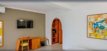 Una habitación con televisión montada en la pared, un escritorio de madera con silla, y un cuadro abstracto de colores en la pared.