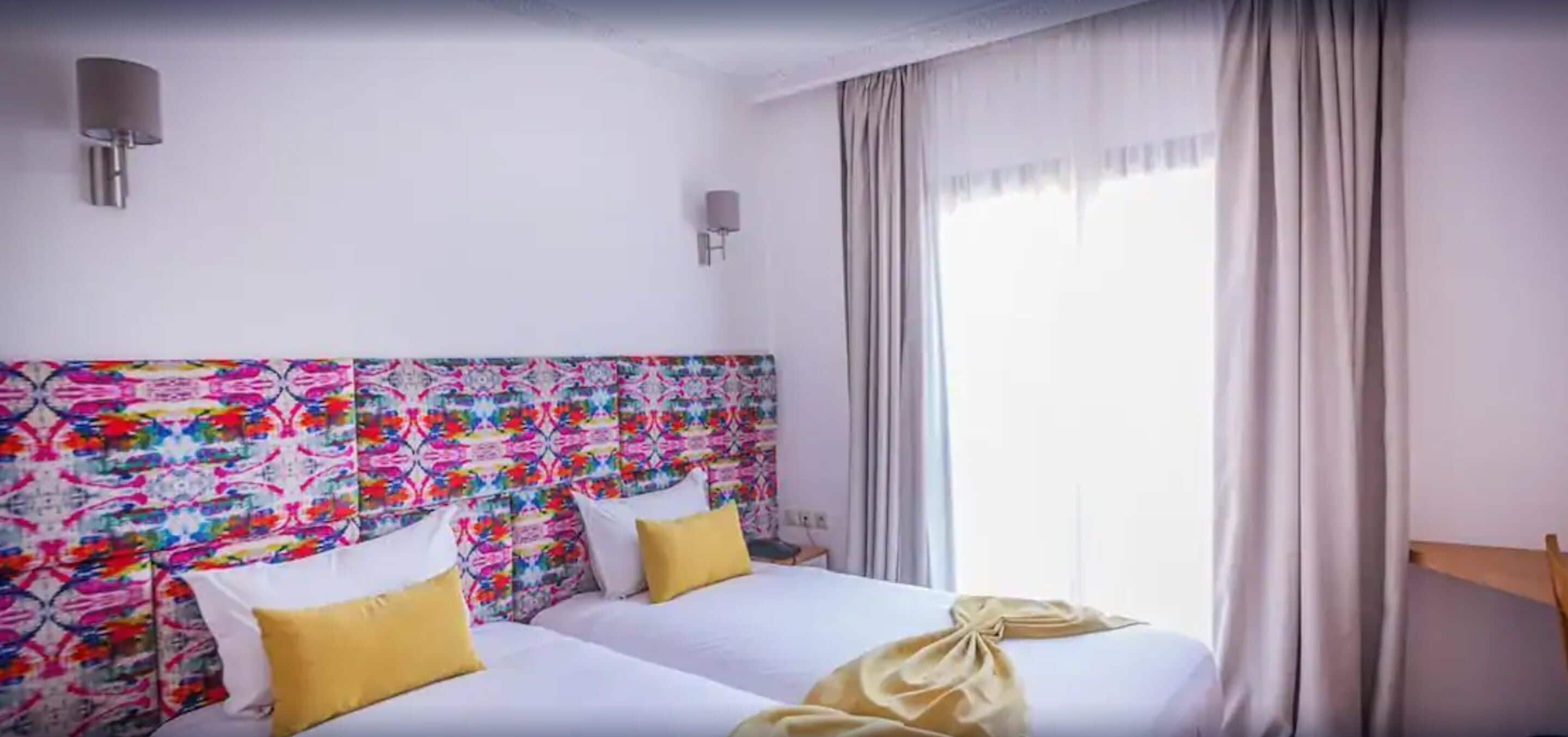 Een hotelkamer met twee bedden, een kleurrijk hoofdbord, witte beddengoed en een groot raam met gordijnen.