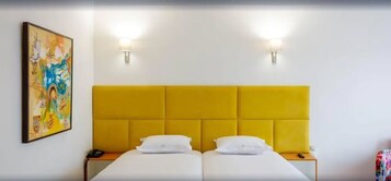 A hotel room with a yellow headboard, white bedding, two bedside tables, and a painting on the wall.