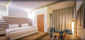 Una camera d'albergo moderna con un letto grande, una testiera in legno, due poltrone, un tavolino e una grande finestra con tende.