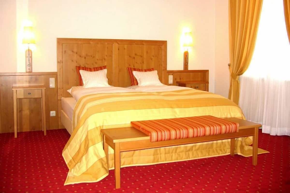 A hotel room with a wooden headboard, two bedside tables with lamps, a bed with striped bedding, a bench, and a red carpet.
